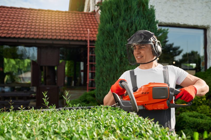 Handsome young man in safety mask and gloves shaping overgrown bushes at garden. Caucasian male using hand electric cutter for work outdoors.